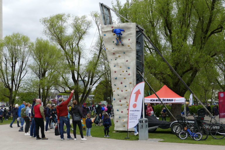 Das Foto zeigt einen Kletterturm an dem gerade ein Kind klettert. Mehrere Leute stehen drumherum und schauen es sich an.