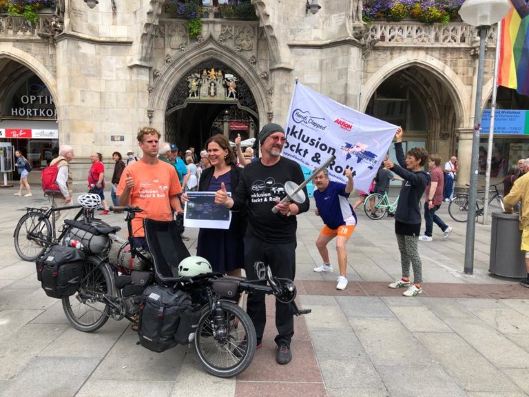 Einige Menschen mit Fahrrädern, Fahnen und einer Fackel demonstrieren auf dem Marienplatz in München. Auf einem Banner steht "Inklusion rockt and rollt".