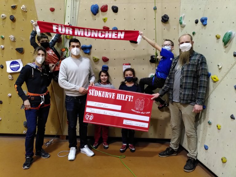 Einige Erwachsene und Kinder stehen und hängen an einer Kletterwand. Sie halten einen roten Schal mit der Aufschrift "Für München" in der Hand sowie einen großen roten Scheck über 5000 Euro mit der Aufschrift "Südkurve hilft."