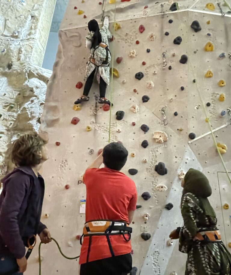 3 Personen, zu sehen von hinten vor einer Kletterwand. Eine weitere Person klettert oben an der Wand und wird von den untenstehenden gesichert.