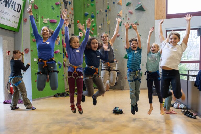 Sieben Kinder beim Luftsprung vor einer Kletterwand