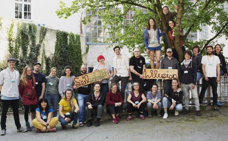 Eine Gruppe on Menschen steht unter einem Baum. einige halten zwei Schilder, auf denen steht "Seilschafft" und "Inklusion"