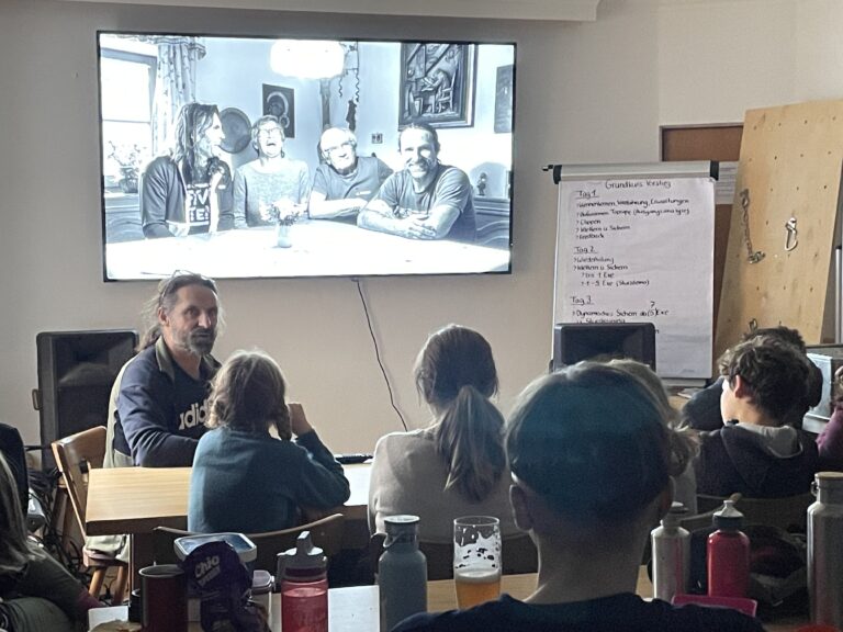 Alexander Huber sitzt vor einer Leinwand, auf das ein Foto projizietr ist. Vor ihm sitzen IWDR Mitglieder und hören seinem Vortrag zu.